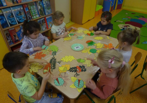 Sześcioro dzieci siedzi przy stole na którym rozłożone są kolorowe liście papierowe oraz owoce. Dzieci dobierają owoce do liści drzew.